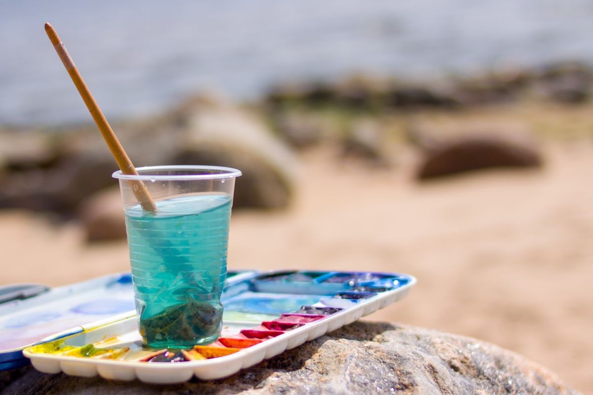 creation, drawing and freedom concept - Artistic equipment: paint brushes, tubes of paint, palette and paintings on rock in nature sea shore at sunny summer day